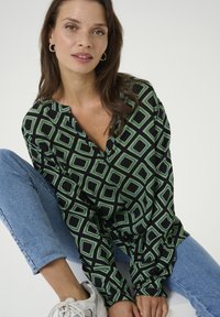 Haut noir et vert à motif avec un design en diamant, à coupe décontractée et col à patte. Porté avec un jean bleu clair.