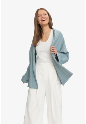 Uśmiechnięta kobieta o długich brązowych włosach, ubrana w biały top, szerokie białe spodnie i luźną niebieską kurtkę, trzymająca klapę kurtki na białym tle.