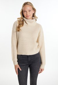 Beżowy sweter robiony na drutach z wysokim kołnierzem typu golf, prążkowanymi mankietami i dołem, z teksturowanym wzorem. Połączony z ciemnymi dżinsami.