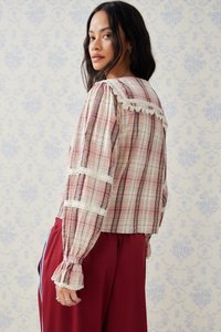 Blusa a quadri rosa con dettagli in pizzo e maniche a sbuffo, caratterizzata da una forma squadrata e un tessuto testurizzato. Abbinata a pantaloni rossi. Sfondo azzurro chiaro.