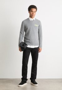 Pilkas megztinis su "FUTURE" logotipu neoninės žalios spalvos, dėvimas ant baltos marškinėlių su apykakle, kartu su juodomis kelnėmis ir juodais sportbačiais.