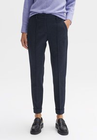 Žena v tmavě modrých slim-fit kalhotách s manžetami, v kombinaci s fialovým svetrem a černými mokasíny, stojí s jednou rukou v kapse.