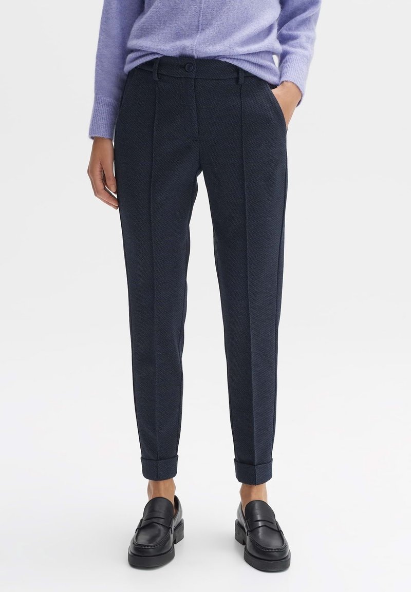 Žena v tmavě modrých slim-fit kalhotách s manžetami, v kombinaci s fialovým svetrem a černými mokasíny, stojí s jednou rukou v kapse.