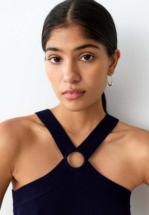 Jeune femme aux cheveux foncés tirés en arrière porte un haut dos-nu bleu marine avec un détail d'anneau circulaire au niveau de l'encolure sur un fond uni.