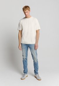 Lichtbeige t-shirt met een ronde hals en korte mouwen; gecombineerd met lichtblauwe jeans en grijze sneakers met beige accenten.