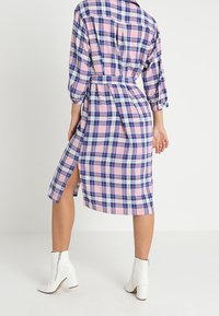 Kvinna som bär en rosa- och blårutig midiklänning med sidospalt och vita ankelboots, stående mot en enfärgad ljus bakgrund.