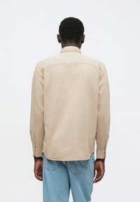 Moški stoji s hrbtom obrnjenim proti kameri, oblečen v bež majico z dolgimi rokavi in svetlo modre kavbojke, pred enobarvno belo kuliso.