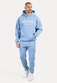 Sudadera azul claro y pantalones de chándal a juego con una textura suave. Presenta el texto "SMILODOX ATHL WEAR" en blanco, bolsillo canguro y puños elásticos.