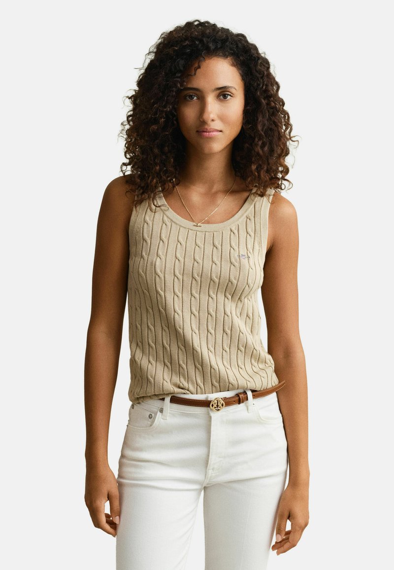 Femme aux cheveux bouclés portant un haut beige en maille torsadée sans manches, un pantalon blanc et une ceinture marron avec une boucle dorée, se tenant devant un fond blanc.