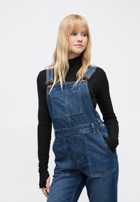 Τζιν σαλοπέτα μεσαίου ντεγκραντέ, με ρυθμιζόμενους ιμάντες, μπροστινή τσέπη, πλαϊνές τσέπες και άνετη γραμμή. Φορεμένη πάνω από μαύρο μακρυμάνικο τοπ.