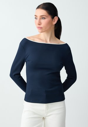 Femme aux cheveux foncés attachés en queue de cheval basse, portant un haut bleu marine à manches longues et épaules dénudées, ainsi qu'un pantalon blanc, regardant sur le côté sur un fond clair.