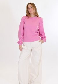 Donna che indossa una maglia rosa a maniche lunghe e pantaloni bianchi a gamba larga, in piedi con una mano nella tasca davanti a uno sfondo bianco semplice.