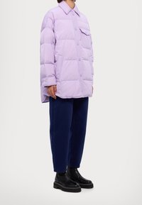 Chaqueta de puffer lavanda con Front con botones, forma oversized, bolsillo frontal y textura suave, combinada con pantalones azul oscuro y botas negras.