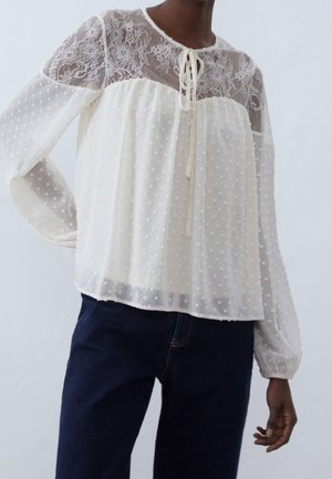 Vrouw draagt een witte doorschijnende blouse met kanten inzetstuk, gestructureerde polkadots, lange mouwen en een strik bij de hals, gecombineerd met donkerblauwe broek.