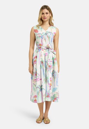 Woman standing wearing a sleeveless, belted midi dress with a colorful palm tree and beach print and gold flat sandals.