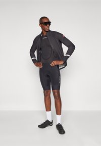 Schwarzes Fahrradoutfit mit einer leichten Jacke und einer Mesh-Unterlage. Die Shorts sind gepolstert und reichen bis zur Mitte des Oberschenkels. Schwarze Schuhe und weiße Socken.