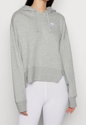 Kobieta ubrana w jasnoszarą bluzę Nike z kapturem i sznurkami oraz białe legginsy, bluza krótka z logo po lewej stronie klatki piersiowej.