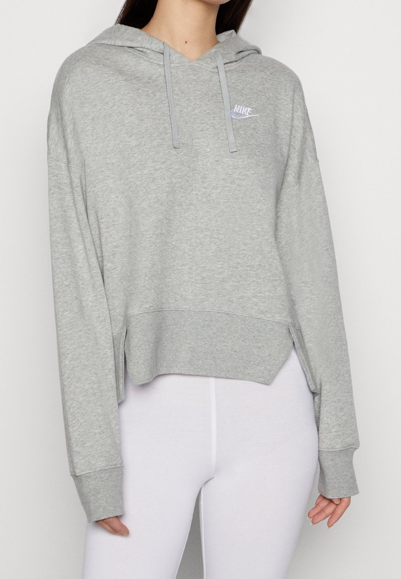 Femme portant un sweat à capuche Nike gris clair avec cordons de serrage et des leggings blancs, sweat court avec logo sur la poitrine gauche.