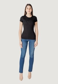 Camiseta negra ajustada con un logo texturizado de "Guess", combinada con unos jeans ajustados de mezclilla azul claro y unos zapatos de tacón nude.