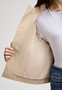 Giacca beige con una texture liscia, caratterizzata da una fodera e cuciture visibili. La parte anteriore aperta rivela una camicia bianca sottostante.