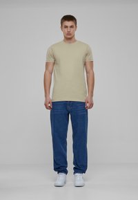 Světle béžové tričko s krátkým rukávem v kombinaci s volnými modrými džíny a bílými teniskami, umístěné na neutrálním pozadí.