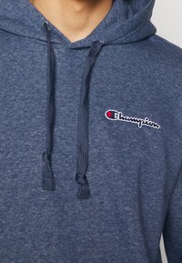 Gros plan sur un sweat à capuche bleu Champion montrant les cordons de serrage et le logo Champion brodé sur la poitrine.