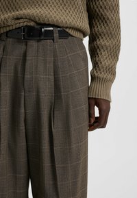 Tan sweater with a textured knit and ribbed cuffs, paired with brown check-pattern trousers. Black belt with a rectangular buckle.