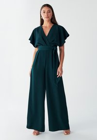 Tumši zaļš ietīšanas kombinezons ar platiem piedurknēm, sasienamu jostasvietu un plašām kāju biksēm; izgatavots no gludas auduma, radot plūstošu siluetu.