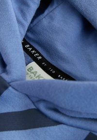 Modra tkanina z mehko teksturo, na kateri je črn etiketa z napisom "TED BAKER" in vidnimi črtastimi dizajnerskimi elementi.