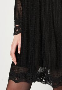 Schwarzes Kleid aus strukturiertem Stoff, mit Spitzenakzenten und einem gewellten Saum. Verfügt über lange Ärmel mit aufwendigen Manschetten und einer lockeren Passform.