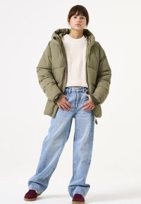 Chaqueta tipo puffer verde olivo con capucha, cierre de cremallera, puños acanalados, combinada con jeans de pierna ancha azul claro y zapatos color granate.