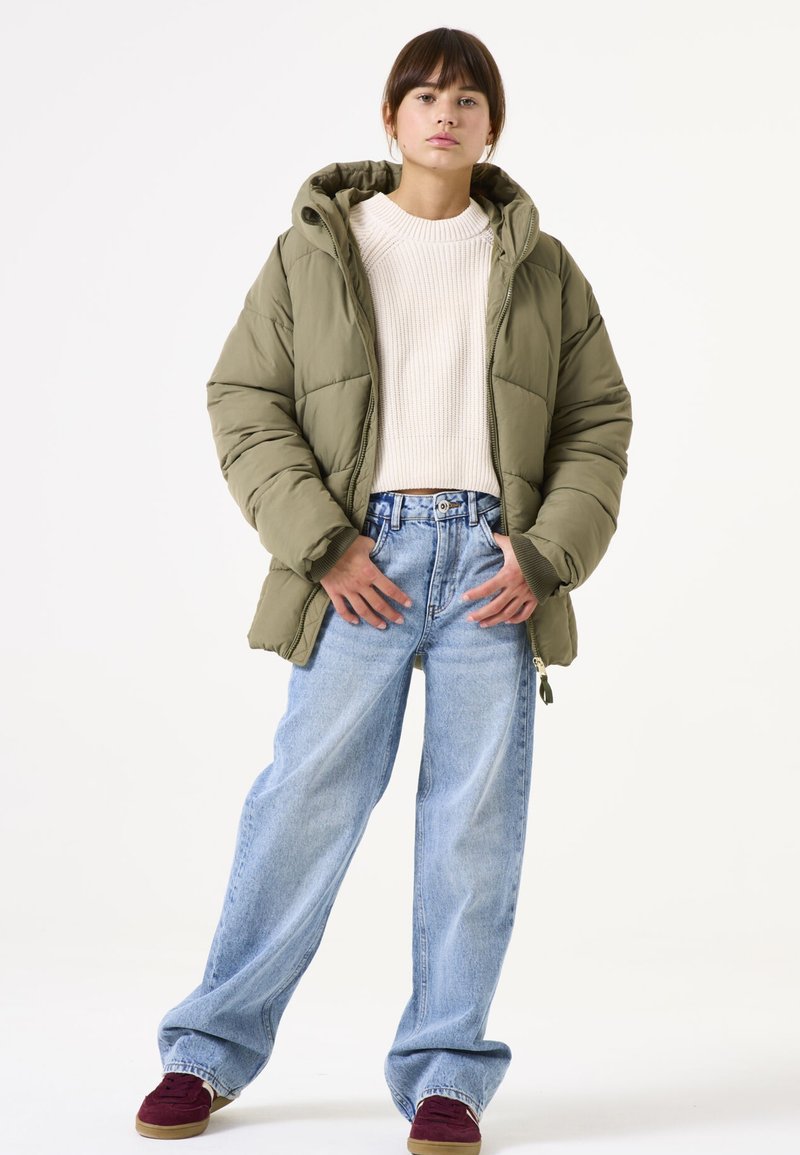 Chaqueta tipo puffer verde olivo con capucha, cierre de cremallera, puños acanalados, combinada con jeans de pierna ancha azul claro y zapatos color granate.