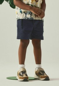 Pantalones cortos azules con cremalleras laterales, combinados con una camiseta de estampado animal multicolor y zapatillas deportivas beige con acentos marrones y calcetines blancos.