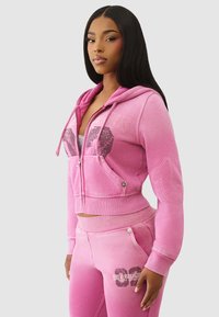Haut à capuche zippé rose avec un design court et des poches avant, associé à un pantalon de survêtement rose avec des graphismes de logo et des détails de couture.