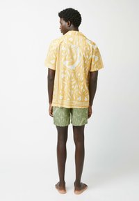 Korte mouwen gele shirt met een wit tropisch patroon, gecombineerd met groene gestreepte shorts, weergegeven vanuit de rug.