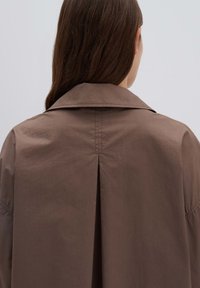 Bruine katoenjas met een brede kraag, rugplooien en rechte snit, met een gladde textuur en minimale details.