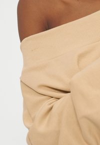 Ορατός γυμνός ώμος και άνω μπράτσο πάνω από μπεζ πλεκτό πουλόβερ χωρίς ώμους με ριπ υφή και πτυχώσεις.