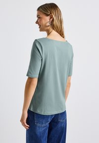Lichtgroene top met korte mouwen en een wijde halslijn, gladde textuur en een ronde zoom. Gepaarde met blauwe high-waisted denim jeans.