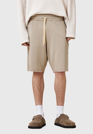 Beige shorts lavet af blødt stof, med en elastisk talje og snøre, samt sidelommer. Parret med brune klipklapper og hvide sokker.
