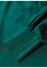 Poignet à manches longues couleur sarcelle avec six boutons recouverts de tissu et un détail froncé sur un tissu lisse, soyeux et léger.