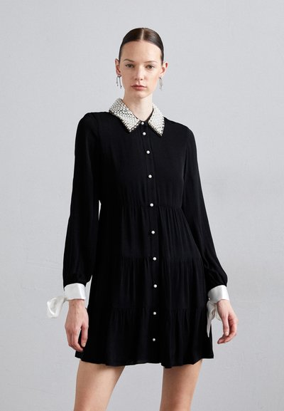 Schwarzes Kleid mit gestuften Design. Hat einen perlenbesetzten Kragen, weiße Knopfdetails und weiße Manschetten mit Bändern. Glattes Material, lange Ärmel.