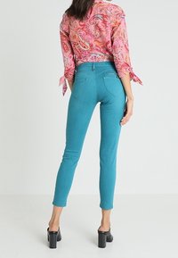 Tyrkysové skinny džíny s vysokým pasem, vyrobené z měkké tkaniny a s minimálním prošíváním. V kombinaci s růžovou halenkou s paisley vzorem a černými lodičkami na vysokém podpatku.