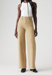 Pantalones de pana khaki de pierna ancha con cintura alta, cierre de botón ajustable, bolsillos frontales y textura suave, combinados con una parte superior ajustada blanca.