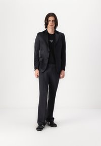 Blazer negro con un acabado texturizado, combinado con una camiseta negra y pantalones oscuros ajustados. Lleva zapatillas negras. Diseño simple y limpio.