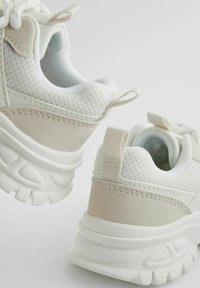 Baskets de sport blanches avec des accents en mesh et en cuir suédé, forme arrondie, semelles en caoutchouc épaisses et languettes au niveau des talons pour un enfilage facile.