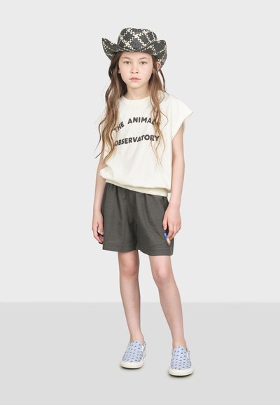 Camisa blanca de manga corta con texto negro, pantalones cortos grises y zapatillas ligeras azul claro con patrones azul oscuro; usando un sombrero de patrón blanco y negro.