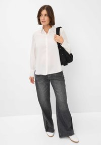 Camicia bianca a maniche lunghe con bottoni, abbinata a jeans larghi color scuro e scarpe bianche, mentre porta una borsa nera. Sfondo neutro.
