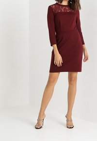 Bordowa sukienka z rękawami o długości trzech czwartych i koronkową górną częścią. Dopasowany krój, gładka tekstura, z długością do kolan. Metaliczne szpilki.