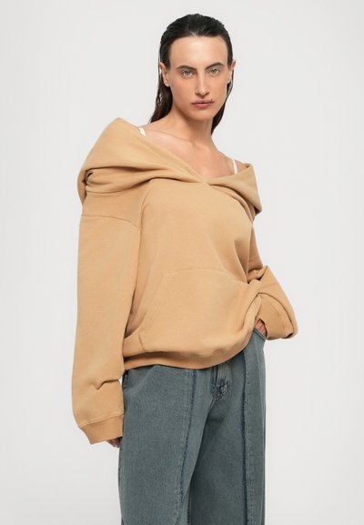 Personne portant un hoodie beige oversize à col off-shoulder et un jean bleu ample, se tenant avec une main dans sa poche sur fond blanc.