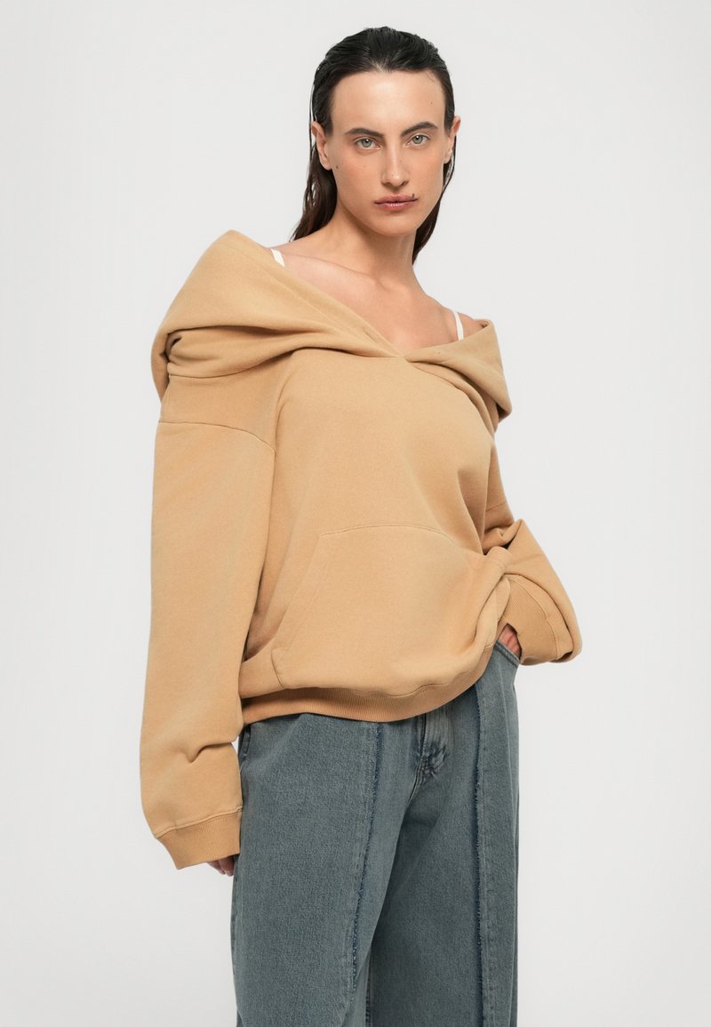 Person som bär en oversized beigefärgad off-shoulder hoodie och lösa blåjeans, står med en hand i fickan mot en vit bakgrund.